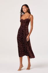 The woman is wearing a sleeveless, dark burgundy dress with thin spaghetti straps. The dress features a fitted bodice with a sweetheart neckline and a floral velvet pattern throughout. It has a midi length, ending below the knees, with a slightly flared hem. She is also wearing gold hoop earrings, a gold ring, and gold high-heeled sandals with thin straps.